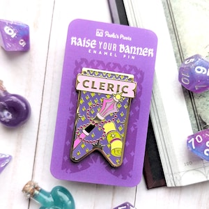 May include: A purple enamel pin with a gold border, featuring a banner with the word "CLERIC" in pink letters. The banner depicts a staff, a scroll, and a string of beads. The pin is on a purple card with the text "Raise Your Banner Enamel Pin" and the logo "Paula's Pixels".