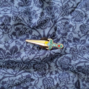 May include: A yellow and blue enamel pin of a dagger with a red gem on the handle. The pin is on a blue fabric with a floral pattern.