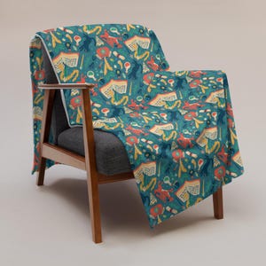 May include: A teal throw blanket draped over a wooden armchair. The blanket features a repeating pattern of open books, mushrooms, and other whimsical illustrations in shades of red, yellow, and blue. The chair has a gray seat and back.