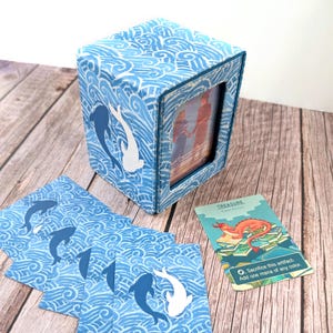 May include: A blue box with a photo frame, featuring a wave pattern and dolphin silhouettes. Several matching cards with dolphin designs are scattered around. A game card with a dragon illustration is also present.