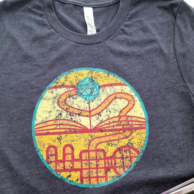 May include: A dark gray t-shirt with a vintage-style circular graphic. The graphic features a blue and yellow dice, a red and orange path, and a yellow and orange grid pattern.