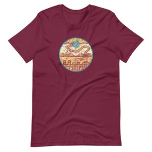May include: A maroon t-shirt with a distressed, circular graphic. The graphic features a book with a winding path leading to a blue and green twenty-sided die.