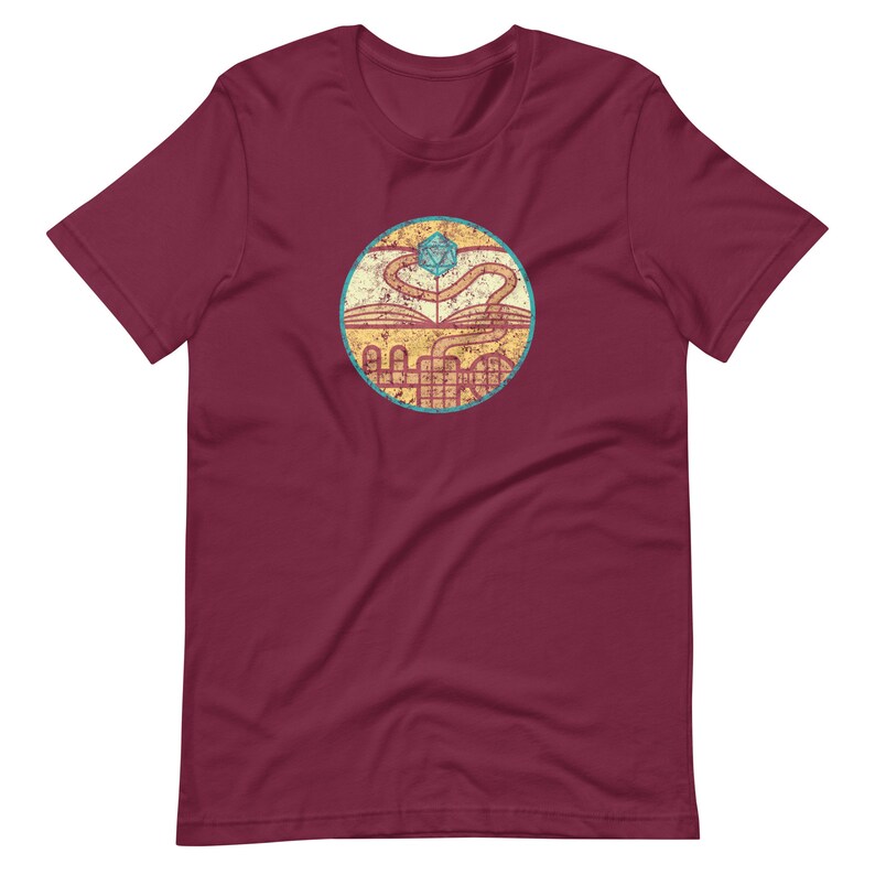 May include: A maroon t-shirt with a distressed, circular graphic. The graphic features a book with a winding path leading to a blue and green twenty-sided die.