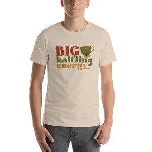 May include: A beige t-shirt with the text "BIG halfling energy" in brown, orange, and green lettering. A green teacup with a brown handle is above the text.