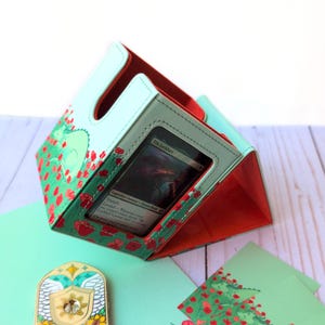 Puede incluir: Un tarjetero verde menta con interior rojo y dise&ntilde;o de flores de amapola. El soporte tiene una ventana transparente que muestra una tarjeta con el texto "Tila Lockbarr". Tambi&eacute;n se ven un peque&ntilde;o alfiler decorativo y una tarjeta a juego.