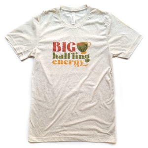 May include: A beige t-shirt with a graphic design that says "BIG halfling energy" with a green teacup illustration.