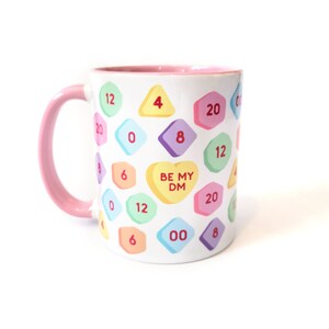 May include: White ceramic mug with a pink rim and handle. The mug has a pattern of colorful candy hearts with numbers and the text "BE MY DM" on a heart.