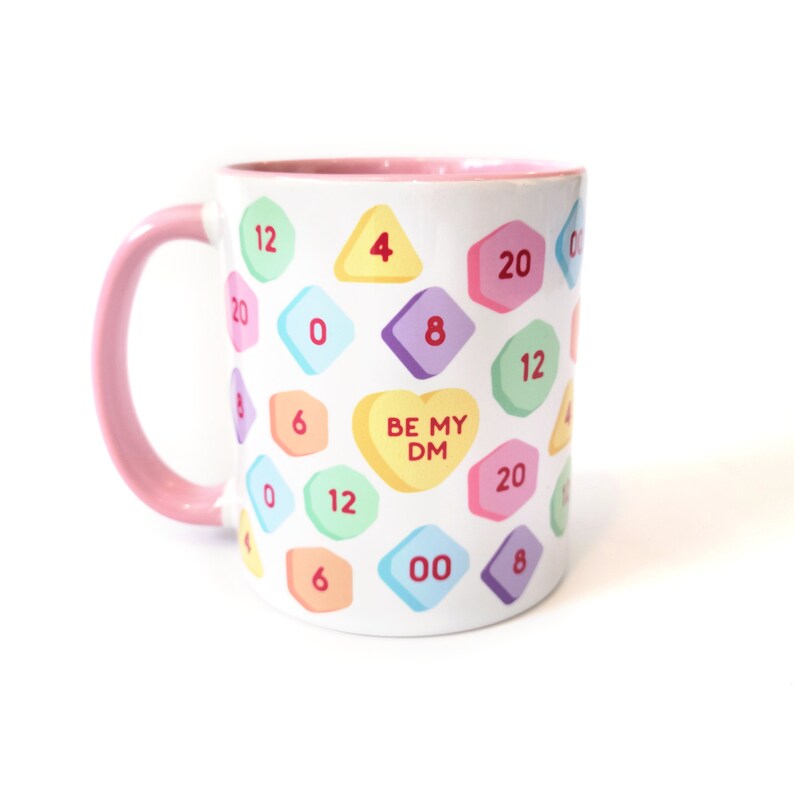 May include: White ceramic mug with a pink rim and handle. The mug has a pattern of colorful candy hearts with numbers and the text "BE MY DM" on a heart.