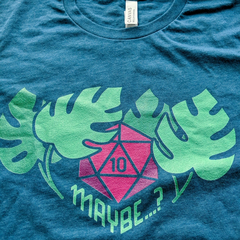 Puede incluir: Una camiseta azul con un dise&ntilde;o verde y rosa. El dise&ntilde;o presenta dos hojas tropicales y un dado rosa y rojo de 10 caras con el texto "MAYBE..." impreso debajo.