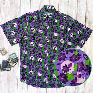 May include: A short-sleeve button-down shirt featuring a repeating pattern of light pink skulls interspersed with dark purple flowers and bright green leaves on a purple background. The shirt has a standard collar and a front button placket.