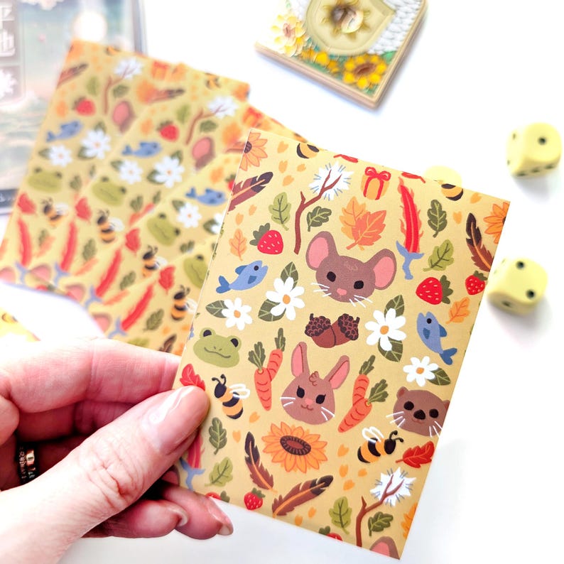 May include: A collection of illustrated stationery featuring a whimsical forest theme. The designs include cartoon animals, flowers, and autumn elements in a warm color palette. The stationery is held by a hand, with dice in the background.