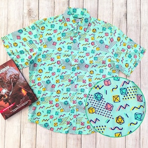 May include: A short-sleeved, light blue button-down shirt with a geometric pattern of dice and abstract shapes in yellow, pink, and teal.