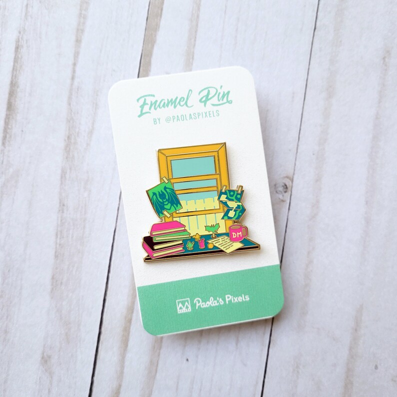 K&ouml;nnte beinhalten: Emaille-Anstecknadel mit einer B&uuml;roszene mit einem Fenster, B&uuml;chern, Pflanzen und einer Tasse mit der Aufschrift "DM". Die Nadel ist golden mit gr&uuml;nen, rosa und gelben Akzenten. Der Text "Enamel Pin" und "By @paolaspixels" befindet sich auf der Karte.