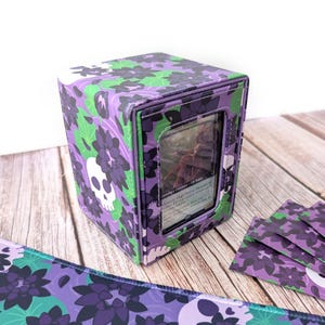 May include: A rectangular deck box featuring a purple and green floral pattern with white skull motifs. The front panel includes a clear window displaying a trading card. The box is constructed with stitched edges and a matching patterned surface.