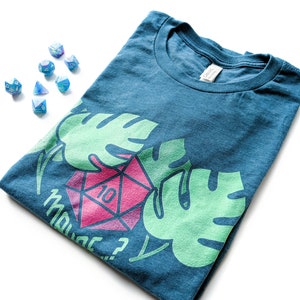 Puede incluir: Una camiseta azul turquesa con un gr&aacute;fico de dados rosa y blanco y hojas tropicales verdes. El texto "How's dice?" est&aacute; impreso en la camiseta.