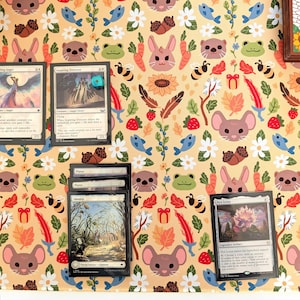 Può includere: Diverse carte di Magic: The Gathering sono esposte su un tappetino da gioco beige con un motivo ripetuto di animali dei cartoni animati, fiori e cibo. Le carte sono in inglese e presentano varie illustrazioni e testo.