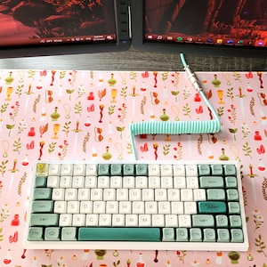 May include: A white and teal mechanical keyboard with a coiled teal cable sits on a pink desk mat with a botanical pattern. The keyboard has white and teal keycaps. The desk mat features illustrations of potions, candles, and leaves.