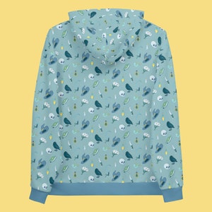 Puede incluir: Una sudadera con capucha azul con un patr&oacute;n repetido de cuervos negros, flores blancas, sombreros azules y otros s&iacute;mbolos de brujer&iacute;a sobre un fondo azul claro.