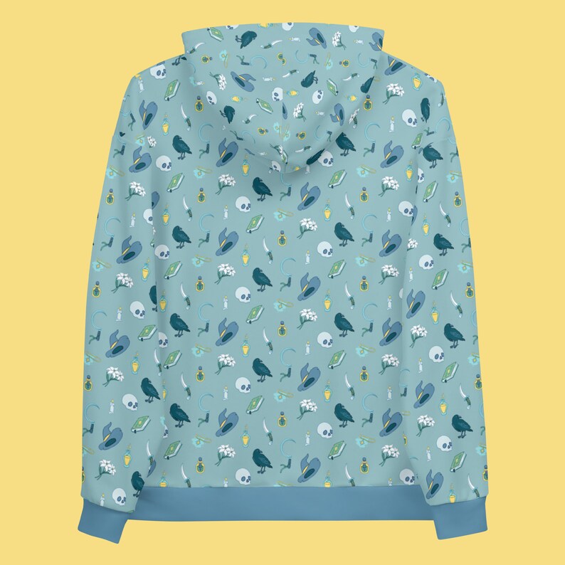Puede incluir: Una sudadera con capucha azul con un patr&oacute;n repetido de cuervos negros, flores blancas, sombreros azules y otros s&iacute;mbolos de brujer&iacute;a sobre un fondo azul claro.