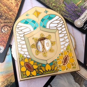 May include: A gold-toned metal enamel pin shaped like an arched window, featuring white wings, yellow sunflowers, and a central shield with a sun emblem. A circular cutout in the center displays the numbers 4 and 0 on a rotating inner dial.