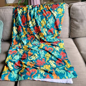 Pu&ograve; includere: Una coperta turchese con un motivo ripetuto di draghi colorati in rosso, giallo, arancione e verde. La coperta &egrave; drappeggiata su un divano grigio, con un lato inferiore bianco e soffice visibile.