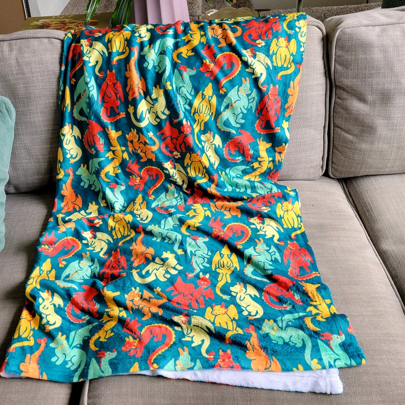 Pu&ograve; includere: Una coperta turchese con un motivo ripetuto di draghi colorati in rosso, giallo, arancione e verde. La coperta &egrave; drappeggiata su un divano grigio, con un lato inferiore bianco e soffice visibile.