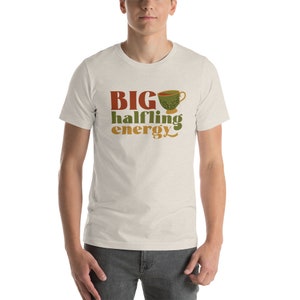 May include: A beige t-shirt with the text "BIG halfling energy" in brown, orange, and green lettering. A green teacup with brown and white details is above the text.