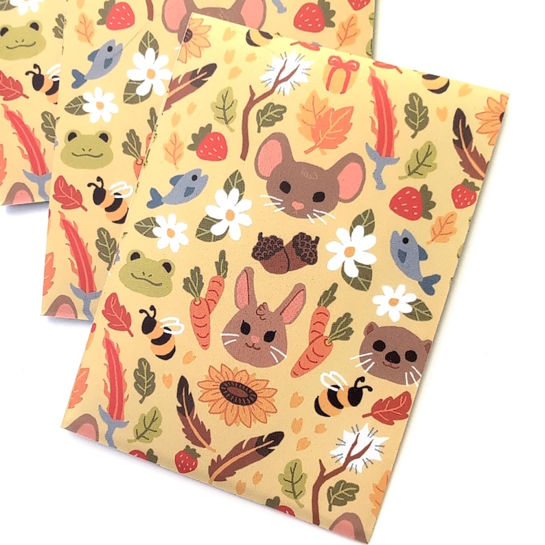 May include: Several envelopes with a repeating pattern of woodland creatures and elements. The design includes a mouse, rabbit, bear, bee, fish, flowers, carrots, strawberries, and feathers on a light yellow background.