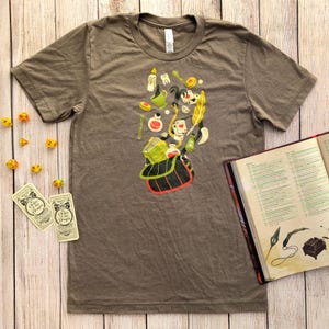 May include: A brown t-shirt with a graphic of a bag spilling out various fantasy-themed items, including a book, potion bottles, and a feather. Dice and cards are scattered around the shirt. A book is open on the right side of the image.