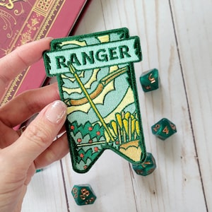 May include: Green felt patch with a green and yellow embroidered banner that says "RANGER". The banner has a design of a bow and arrow with a green, yellow, and orange colour scheme.