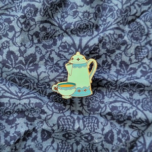 May include: A gold enamel pin featuring a blue and green teapot and a matching teacup. The teapot has a white trim and a cross design. The teacup has a yellow interior.