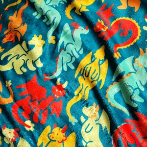 Pu&ograve; includere: Una coperta turchese con un motivo ripetuto di draghi dei cartoni animati colorati. I draghi sono rossi, gialli, arancioni e turchesi. La coperta ha una consistenza morbida e soffice.