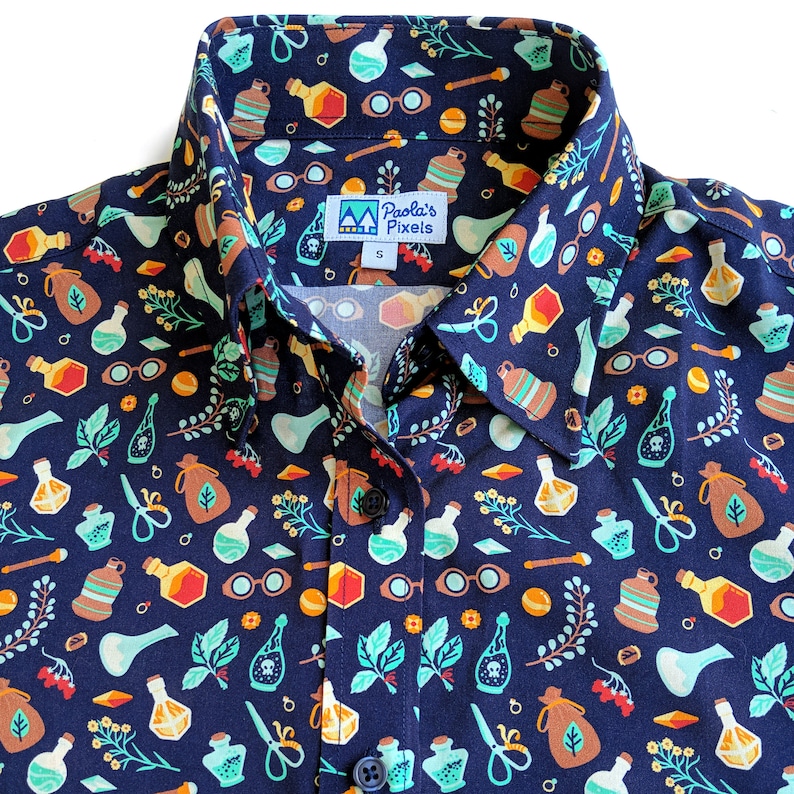 Puede incluir: Una camisa azul marino de botones con un patr&oacute;n caprichoso de frascos de pociones, hierbas y otros elementos m&aacute;gicos. La camisa tiene cuello y un frente abotonado. La etiqueta de la camisa dice "Paola's Pixels".