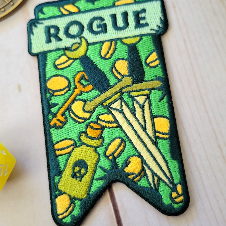 Pu&ograve; includere: Toppa in feltro verde con uno stendardo verde su cui &egrave; scritto "ROGUE" in lettere gialle. Lo stendardo &egrave; decorato con una chiave, un pugnale e una boccetta di pozione, tutto in giallo e verde.