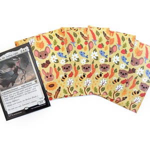 May include: A Magic: The Gathering card and several card sleeves. The card features a black and white illustration of a winged figure. The sleeves have a yellow background with a repeating pattern of cartoon animals, flowers, and other natural elements.