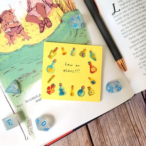 May include: A yellow sticky note with the text "Low on elixirs!!!" surrounded by small cartoon images of potion bottles. The note is on a table with several blue dice.