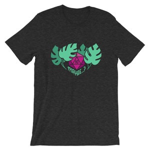 Puede incluir: Camiseta gris oscuro jaspeada con un dado rosa y verde con el n&uacute;mero 10 visible. El dado est&aacute; rodeado de hojas verdes de monstera y el texto "MAYBE...?" est&aacute; debajo del dado.