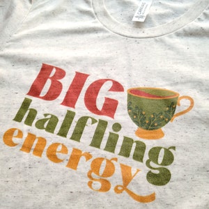 May include: A white t-shirt with the text "BIG halfling energy" in red, green, and orange. A green teacup with a floral design is next to the text.