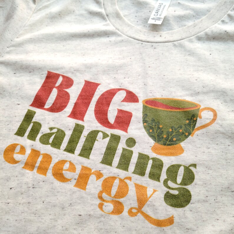 May include: A white t-shirt with the text "BIG halfling energy" in red, green, and orange. A green teacup with a floral design is next to the text.