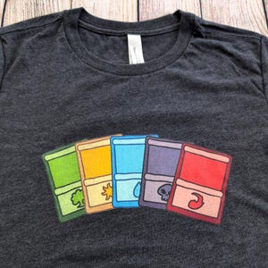 May include: Dark gray t-shirt featuring a colorful graphic of five stylized card designs. Each card is a different color: green, orange, yellow, blue, purple, and red. The cards have unique symbols.