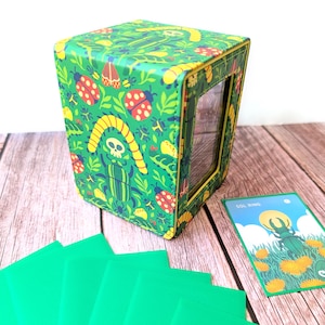 May include: A green box with a colourful insect pattern and a clear window. The box is displayed with green card sleeves and a card with the text "SOL RING" and an insect illustration. The box is on a wooden surface.