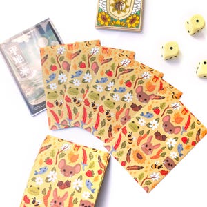 May include: A collection of illustrated stationery featuring a repeating pattern of woodland creatures, including mice, rabbits, bees, and fish, set against a yellow background. Additional items include dice, a wooden box, and a clear plastic case with Japanese text.