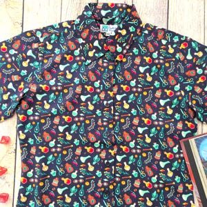 Puede incluir: Una camisa de botones de manga corta azul oscuro con un patr&oacute;n colorido de frascos de pociones, hierbas y otros objetos m&aacute;gicos. La camisa tiene una peque&ntilde;a etiqueta con el texto "Pandas & Pirates".