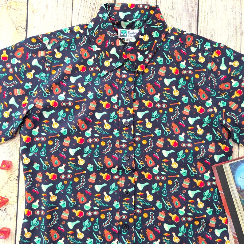 Puede incluir: Una camisa de botones de manga corta azul oscuro con un patr&oacute;n colorido de frascos de pociones, hierbas y otros objetos m&aacute;gicos. La camisa tiene una peque&ntilde;a etiqueta con el texto "Pandas & Pirates".