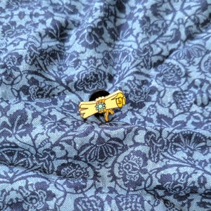 May include: A yellow and blue enamel pin of a rolled up scroll with a blue ribbon and a flower.