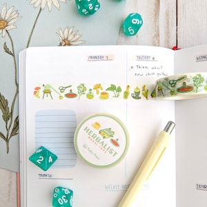 K&ouml;nnte beinhalten: Eine Rolle gr&uuml;nes und wei&szlig;es Washi-Tape mit einem floralen und Zaubertrank-Design. Das Band ist mit "Herbalist" und "Washi Tape" beschriftet, darunter der Text "Pal's Pixels". Das Band befindet sich auf einer wei&szlig;en Seite mit einem gr&uuml;nen Stift und einem gr&uuml;nen W&uuml;rfel.