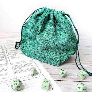 May include: A teal drawstring dice bag with a repeating pattern of stylized creatures and leaves. The bag is made of fabric and has a matching teal drawstring. Several pale green dice and a game sheet are scattered around the bag.
