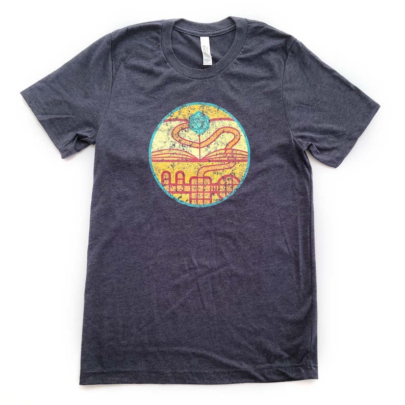 May include: A dark heather grey t-shirt with a vintage-style graphic of a book, a dice, and a winding path. The graphic is in shades of yellow, orange, red, and blue.