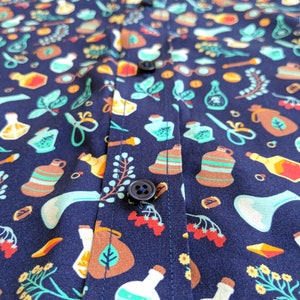 Puede incluir: Una camisa azul marino con un patr&oacute;n repetido de botellas de varios colores, hierbas y otros elementos bot&aacute;nicos. La camisa tiene cierre de botones y un bot&oacute;n en la parte delantera.