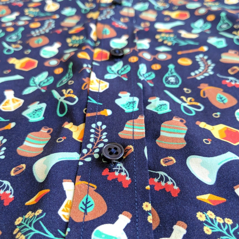 Puede incluir: Una camisa azul marino con un patr&oacute;n repetido de botellas de varios colores, hierbas y otros elementos bot&aacute;nicos. La camisa tiene cierre de botones y un bot&oacute;n en la parte delantera.
